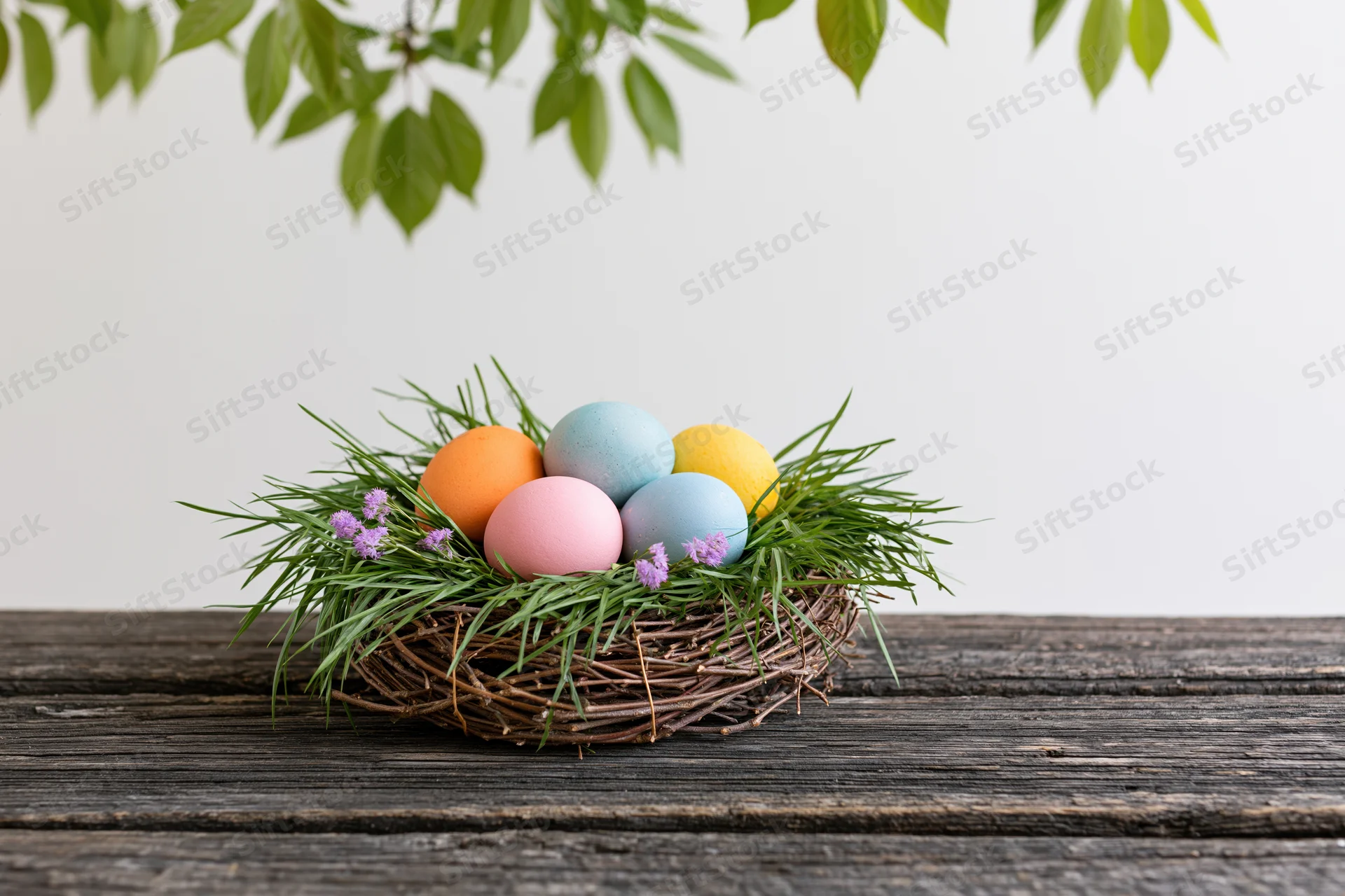 Colorful painted easter eggs in a twig nest with grass and purple flowers on a wooden table under green leaves