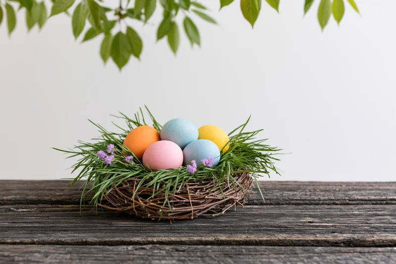 Colorful painted easter eggs in a twig nest with grass and purple flowers on a wooden table under green leaves