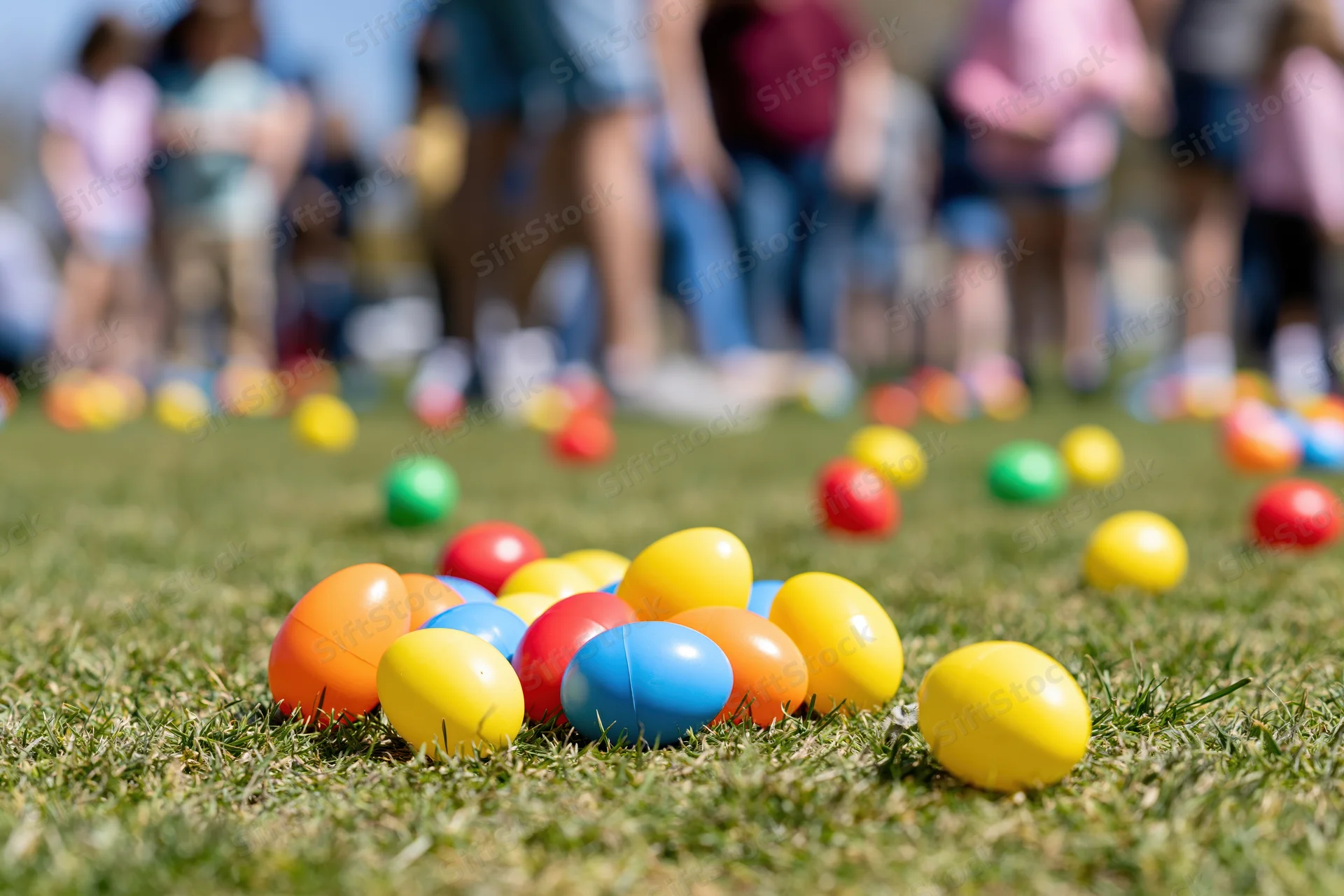 Colorful plastic easter eggs scattered across a grass lawn with children and families in the background during an outdoor spring holiday event