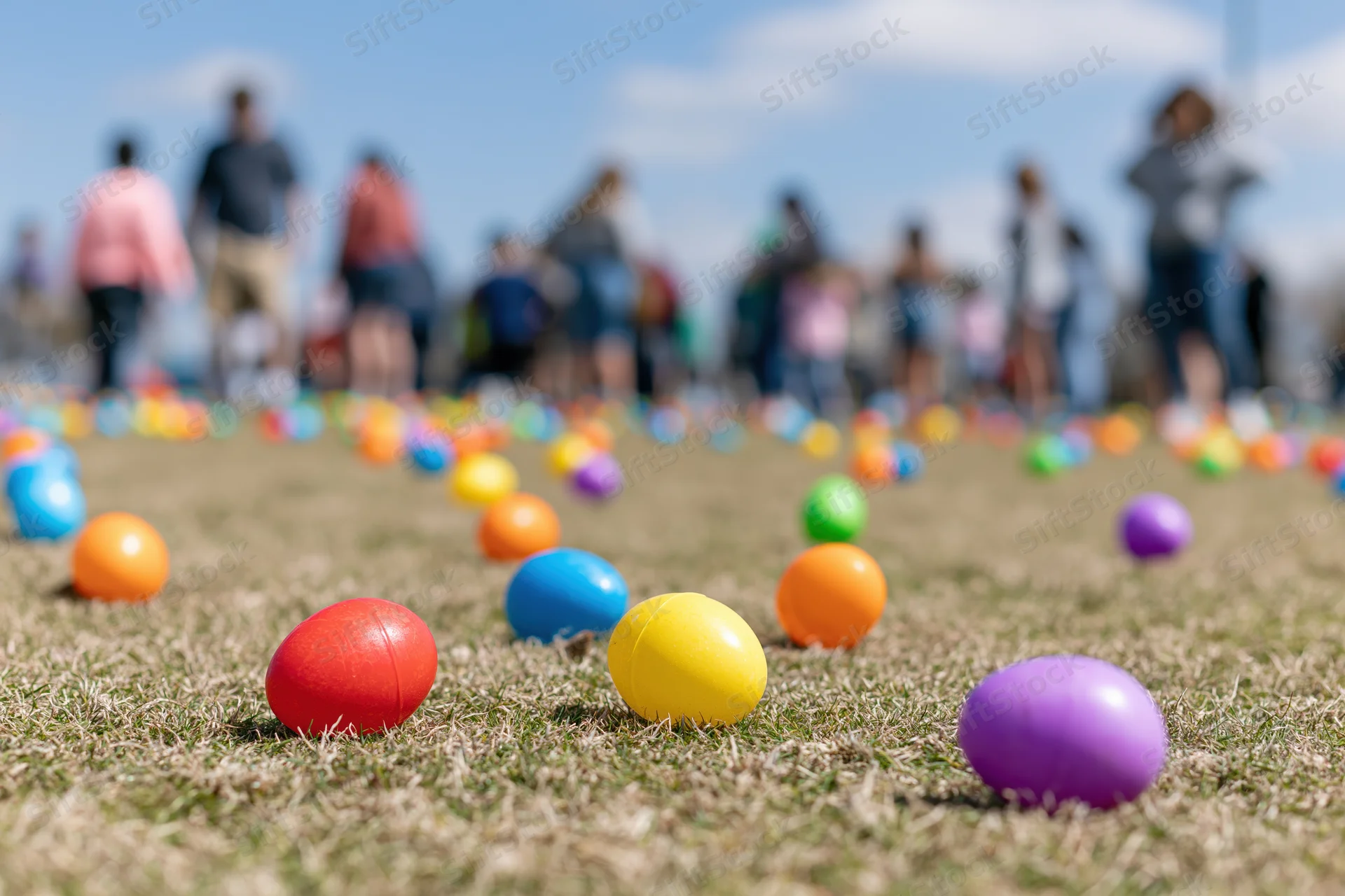 Colorful plastic easter eggs scattered across a mowed grass field with people in the background during a spring holiday event