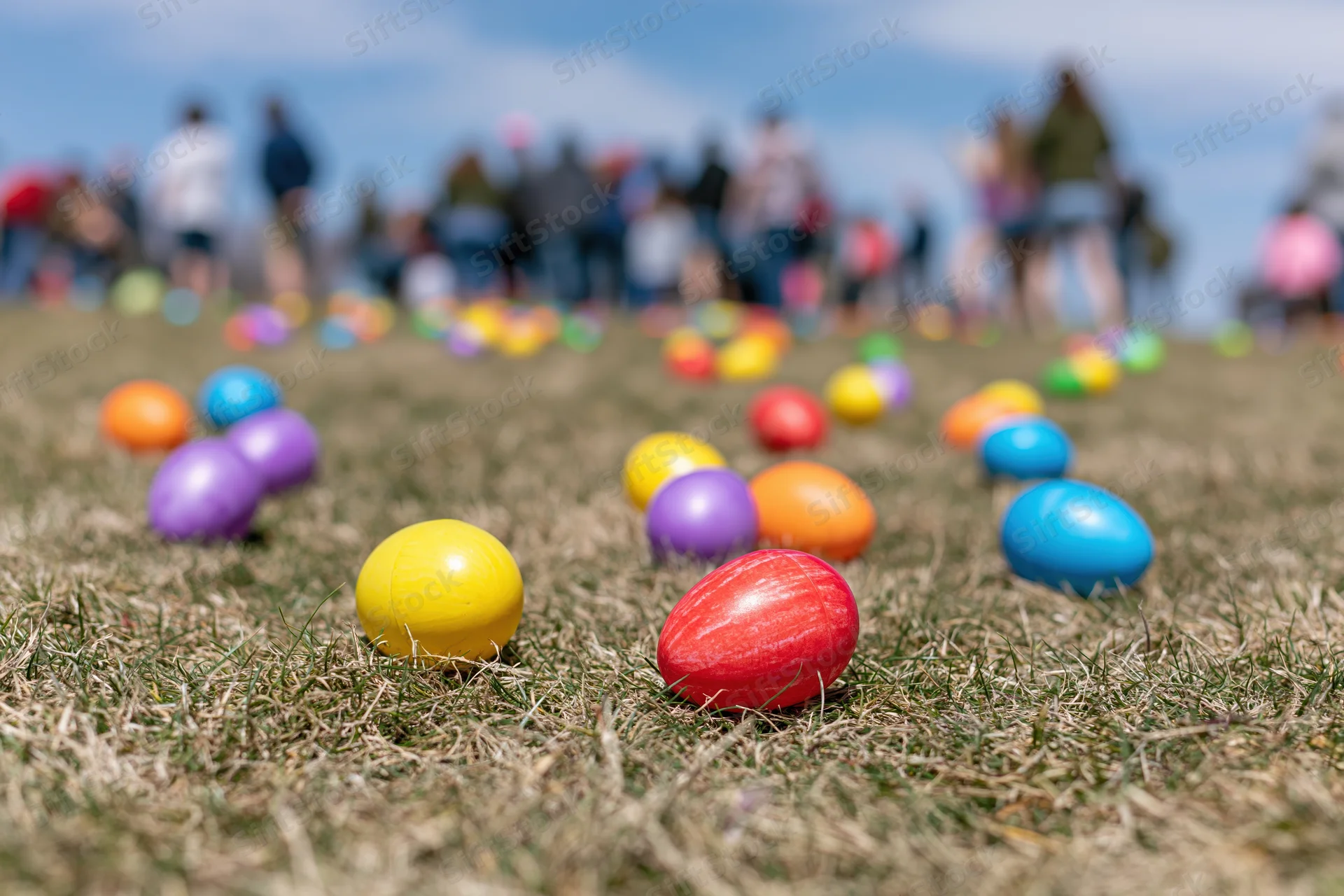 Colorful plastic easter eggs scattered across a mowed grass field with blurred people in the background