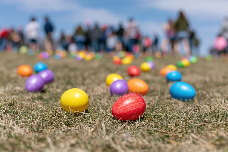Colorful plastic easter eggs scattered across a mowed grass field with blurred people in the background