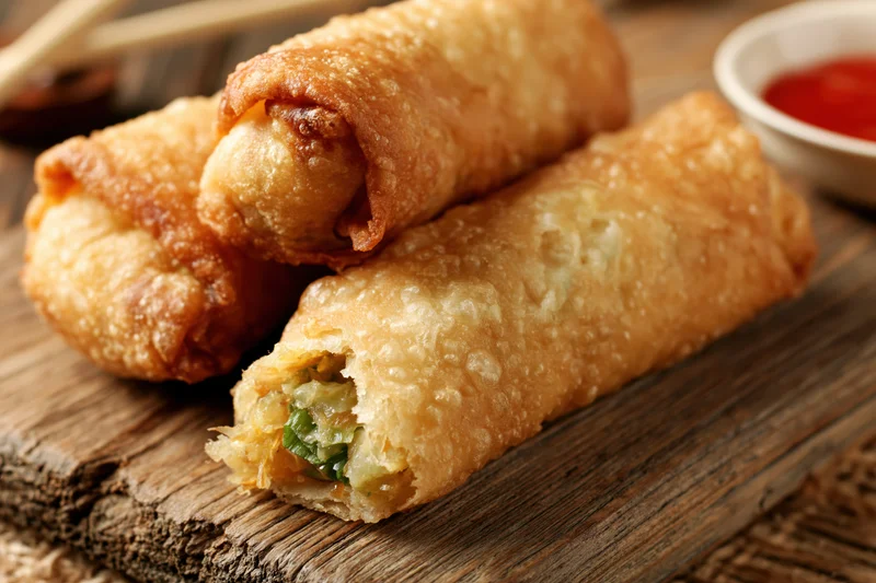 Crispy golden fried egg rolls stacked on a worn wooden cutting board with dipping sauce nearby