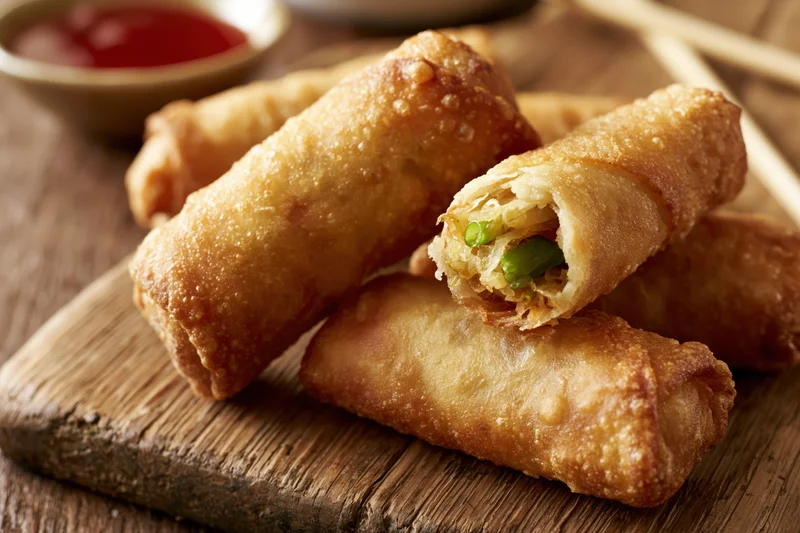 Crispy golden fried egg rolls stacked on a worn wooden cutting board with dipping sauce and chopsticks nearby