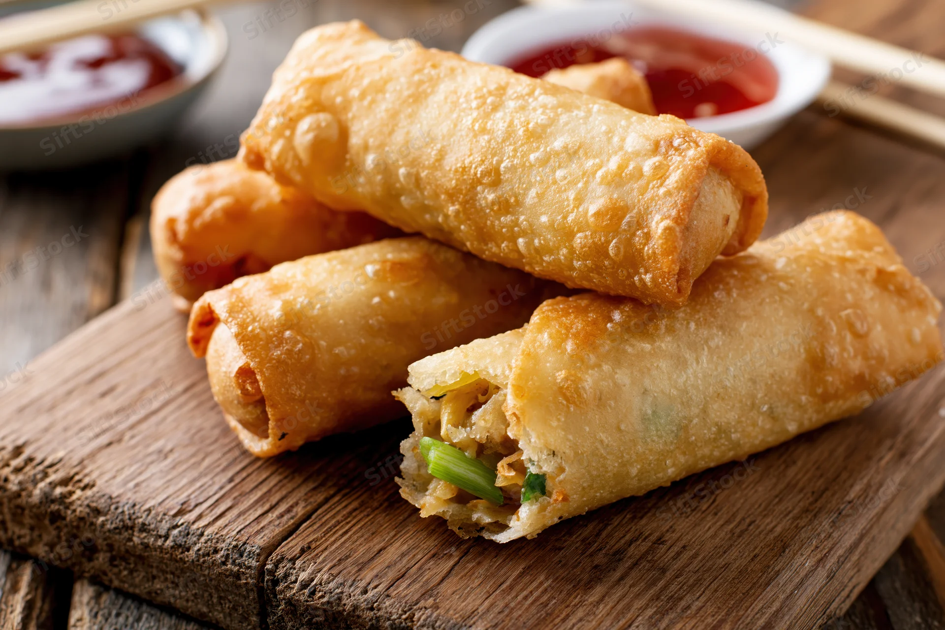 Crispy golden fried egg rolls stacked on a worn wooden cutting board with dipping sauce and chopsticks nearby