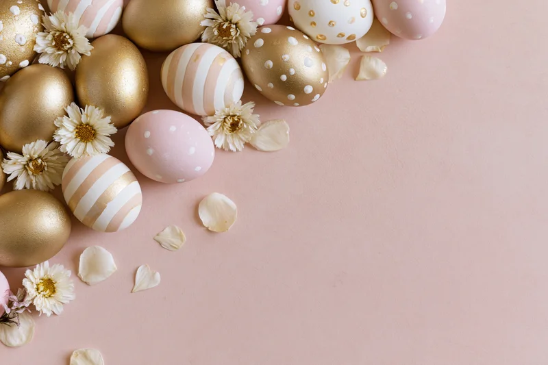 Decorated easter eggs and small white daisies arranged on a pink surface with scattered petals and copyspace