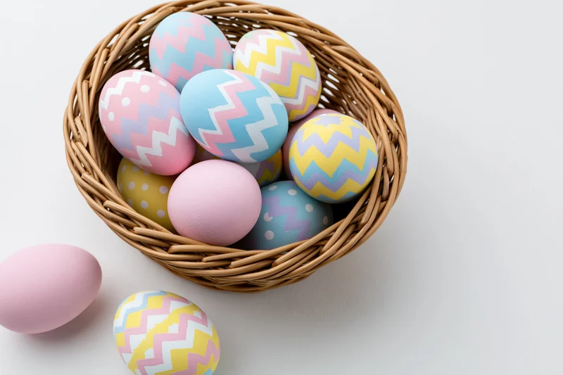 Decorated easter eggs in a woven wicker basket with some scattered beside it on a white surface