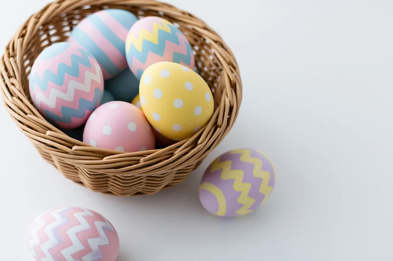Decorated easter eggs in a woven wicker basket with some scattered on white surface