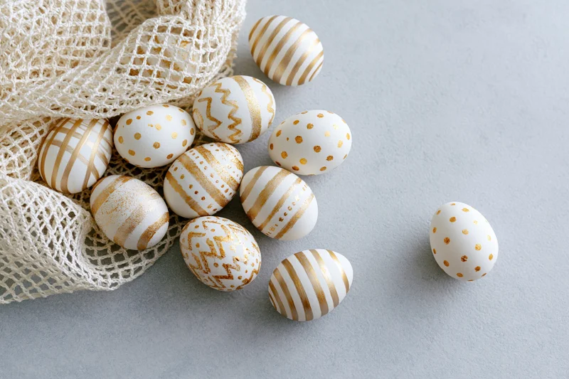Decorated easter eggs with white and gold patterns arranged on a gray surface with a mesh bag