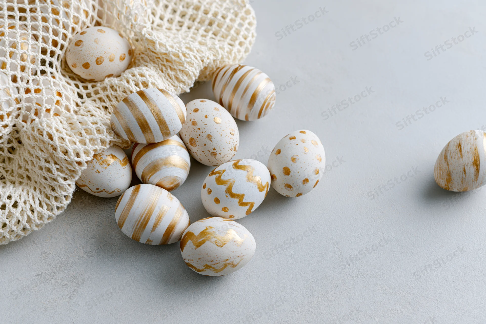 Decorated easter eggs with white and gold patterns scattered on a light surface near a mesh bag