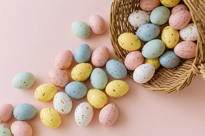 Decorated easter eggs spilling from a tipped wicker basket onto a pastel pink surface