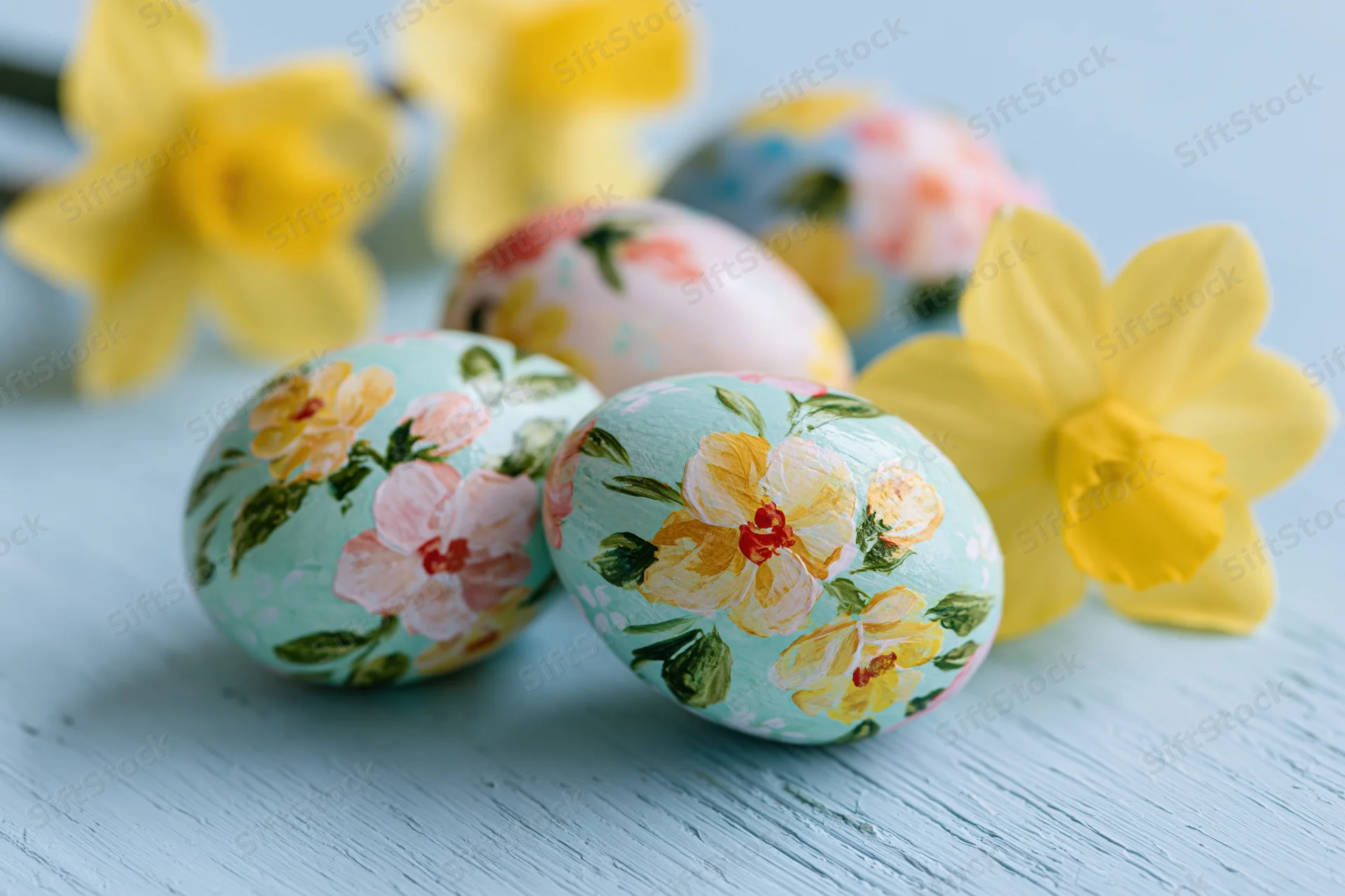 Hand-painted floral easter eggs arranged with yellow daffodils on a light blue wooden surface
