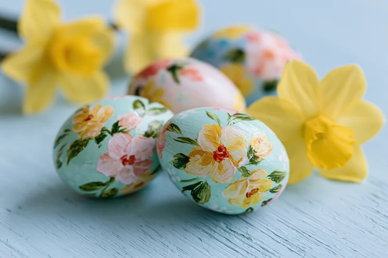Hand-painted floral easter eggs arranged with yellow daffodils on a light blue wooden surface