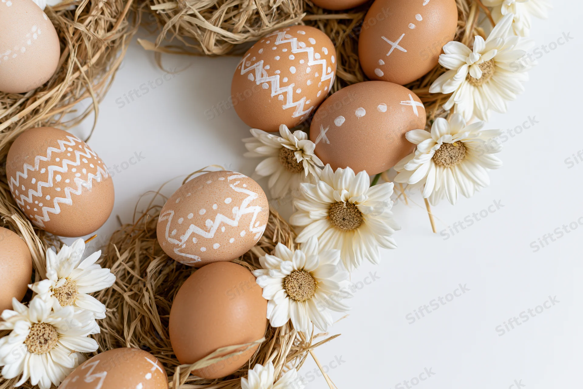 Decorated easter eggs with handdrawn white patterns arranged in straw nests with white daisies on white background