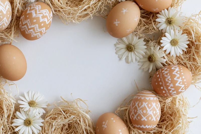 Decorated easter eggs with handdrawn white patterns arranged in a straw frame with daisies on white background