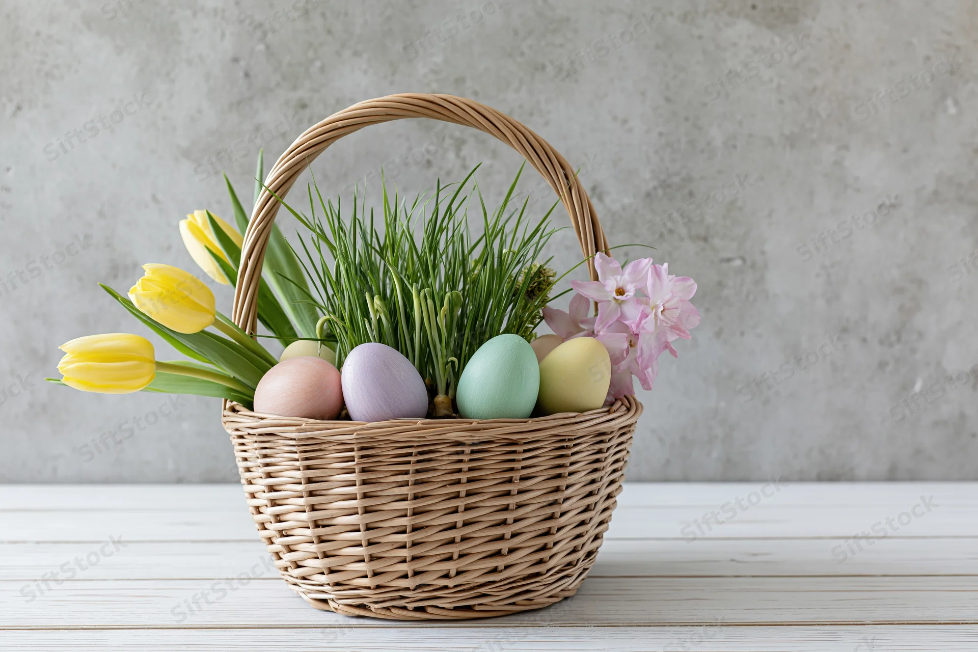 Wicker basket filled with pastel eggs, fresh green grass, yellow tulips and pink flowers on a white wooden table