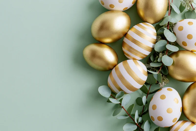 Decorated easter eggs with gold stripes and polka dots arranged with eucalyptus leaves on a mint green surface