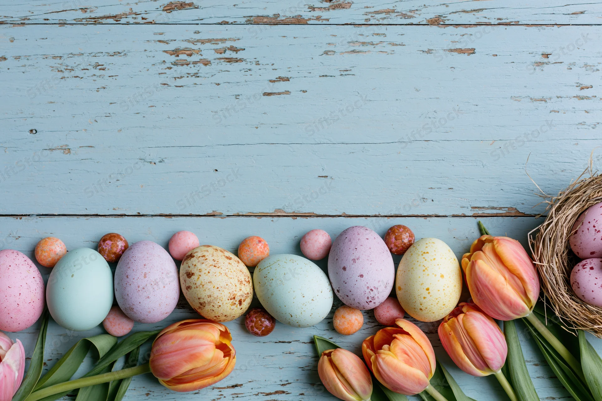 Pastel easter eggs and tulips arranged on a blue wooden surface with a nest and chocolate candies