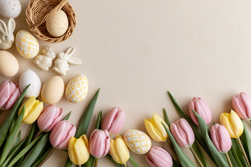Easter flat lay with tulips, eggs, bunnies and basket arranged along bottom edge of cream surface