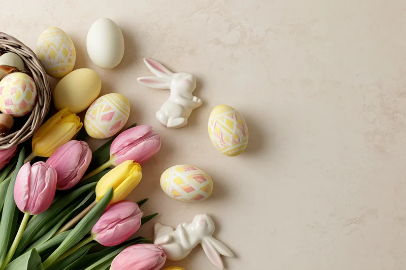 Easter flat lay with tulips, decorated eggs, ceramic bunnies and a basket arranged along the bottom edge of a cream surface