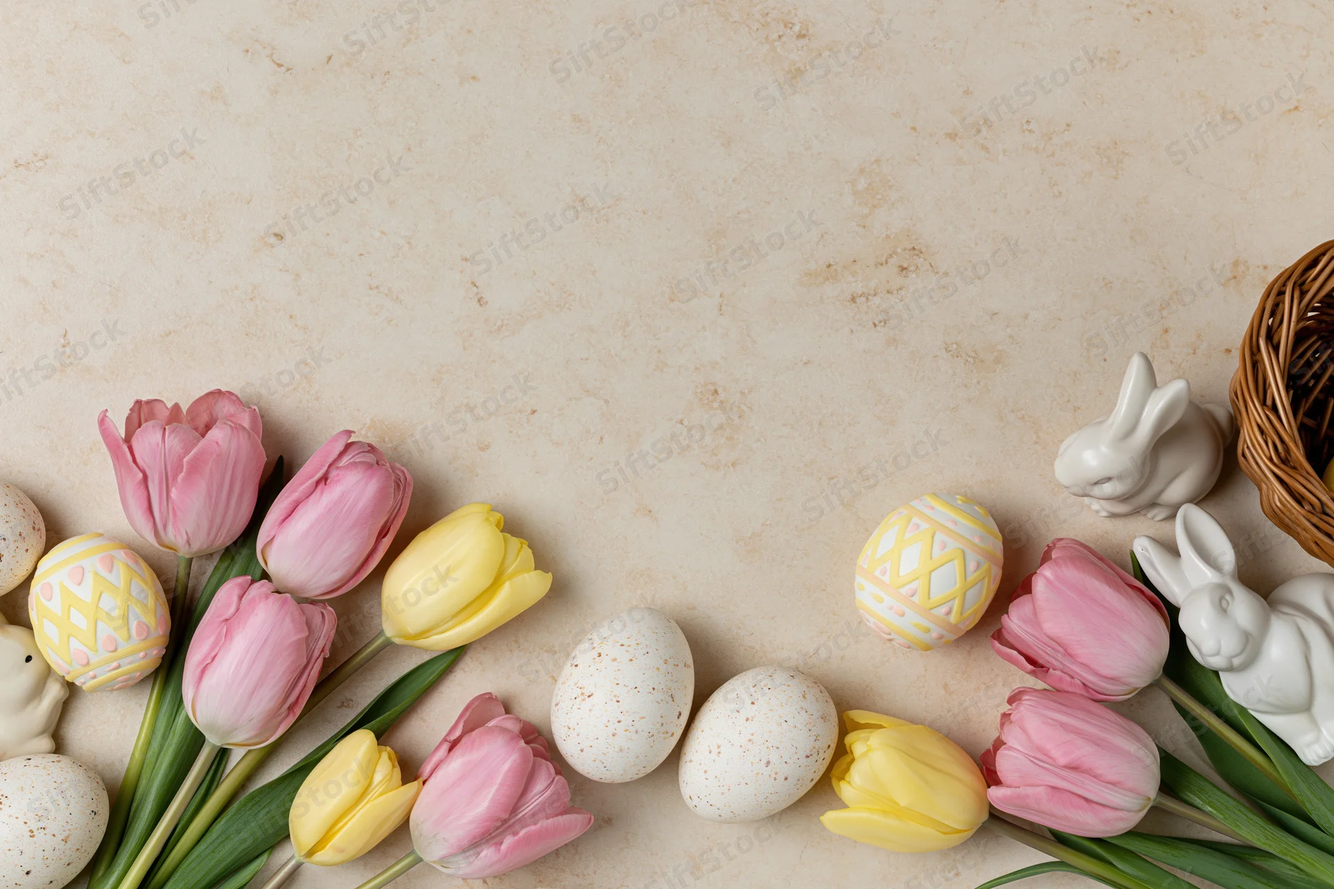 Easter flat lay with tulips, decorated eggs, ceramic bunnies and a wicker basket arranged along the bottom edge of a cream surface