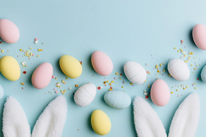 Pastel decorated eggs and bunny ears arranged flat on blue surface with confetti and copyspace