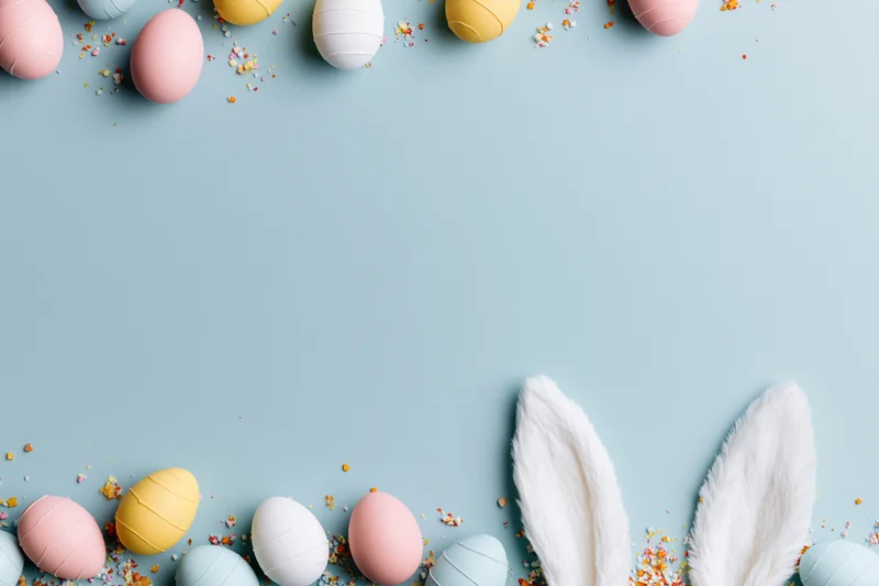Pastel-colored easter eggs and bunny ears arranged in flat lay on light blue background with confetti and copyspace