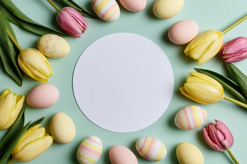 Easter flat lay with yellow and pink tulips and decorated eggs arranged around a white circular mockup on a pastel green background