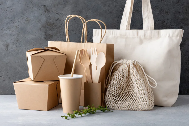 Eco-friendly packaging items arranged on a light gray surface with reusable bags and wooden cutlery