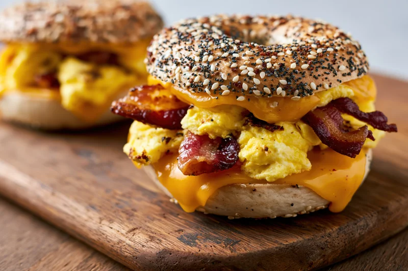 Everything bagel breakfast sandwich with scrambled eggs, bacon, and melted cheese on a wooden board