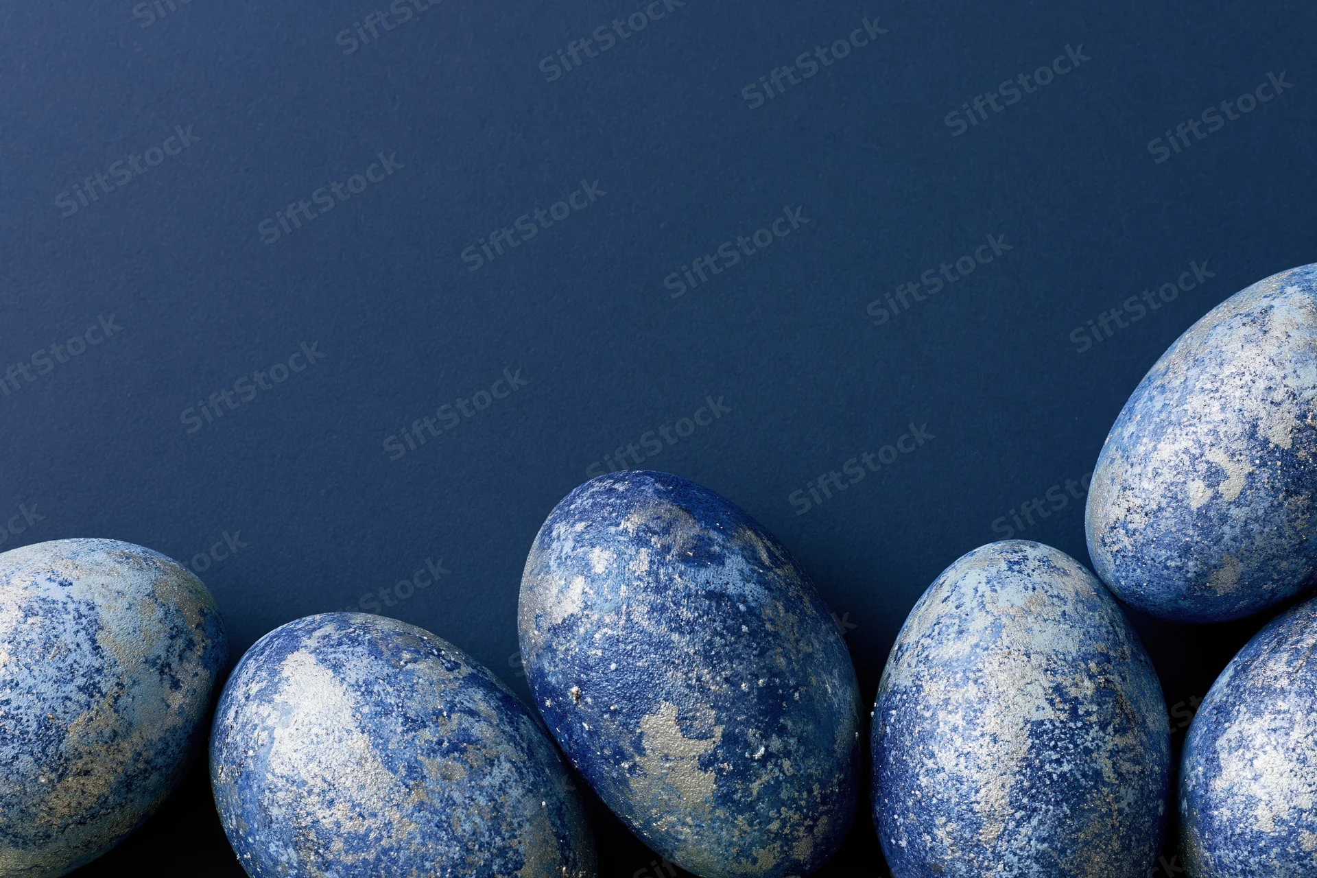 Five blue-painted easter eggs with mottled silver and white finish arranged in a row on a dark blue background with copyspace above