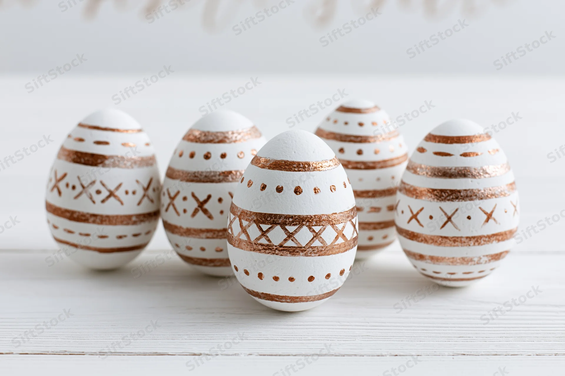 Five white easter eggs with hand-painted copper geometric patterns arranged on a white wooden surface