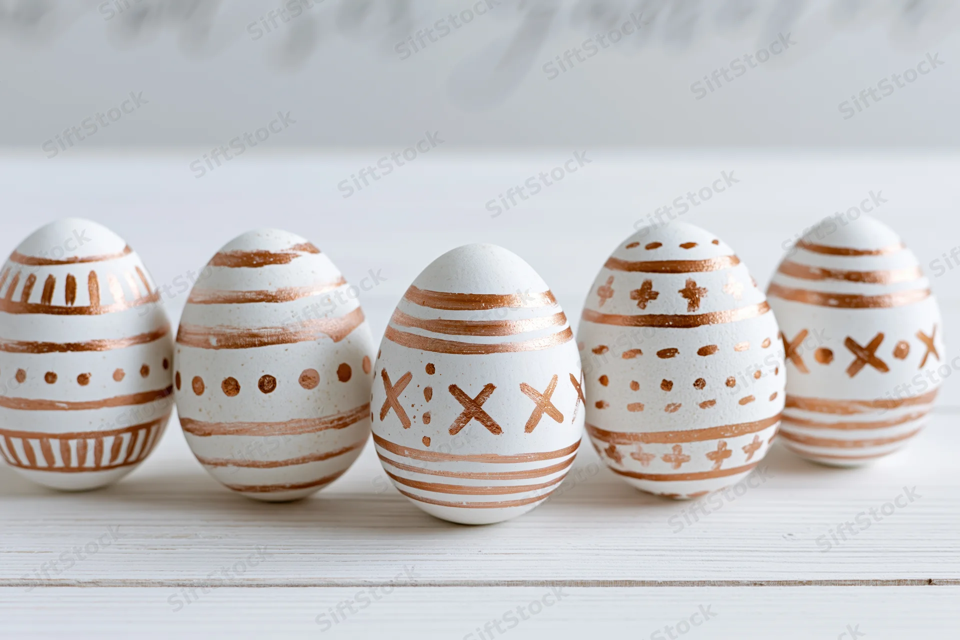 Five white easter eggs with hand-painted copper geometric patterns arranged in a row on a wooden surface