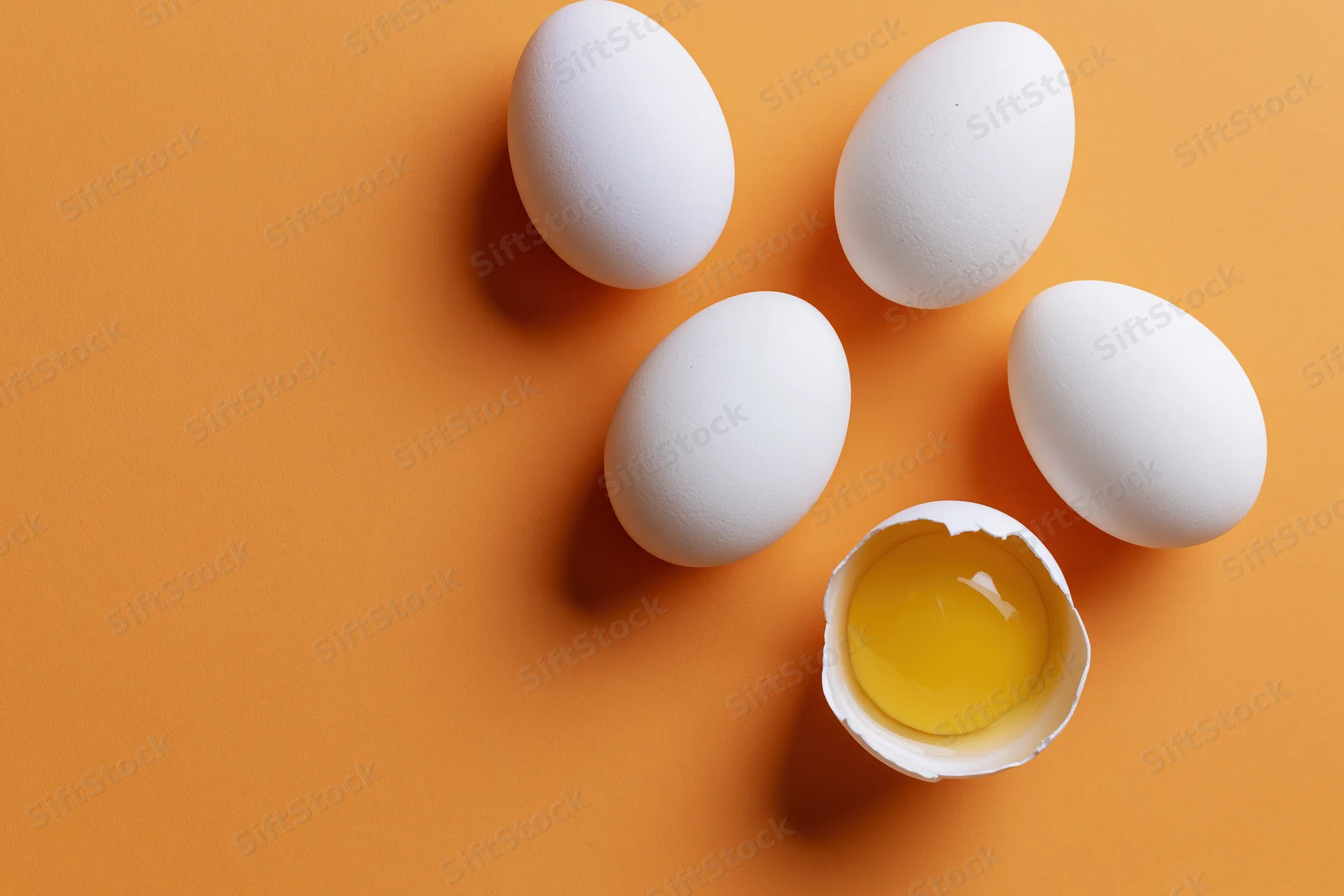 Five white eggs scattered on orange surface with one cracked open revealing yolk