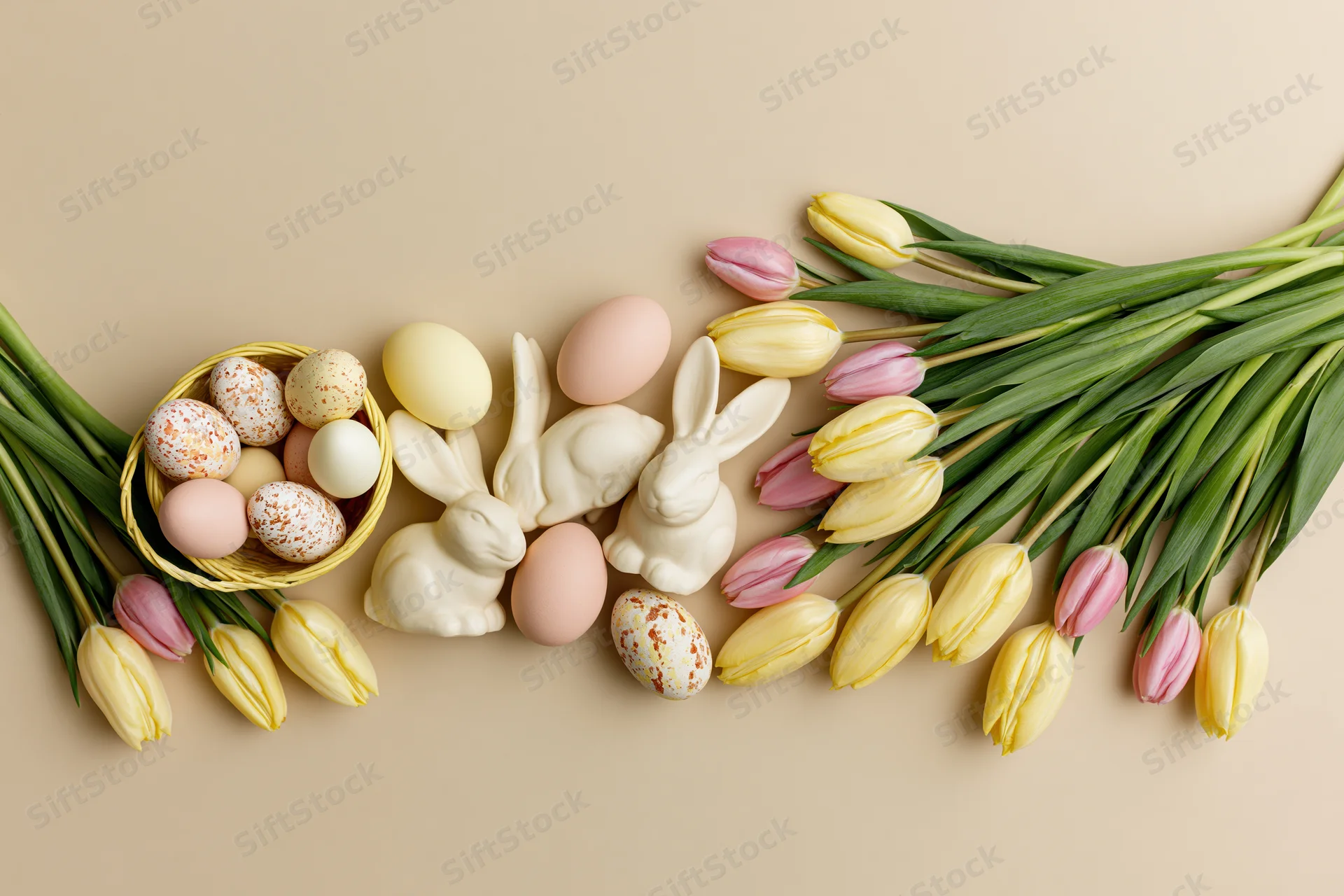 Easter flat lay with tulips, eggs, and bunnies arranged along the bottom edge of a beige surface