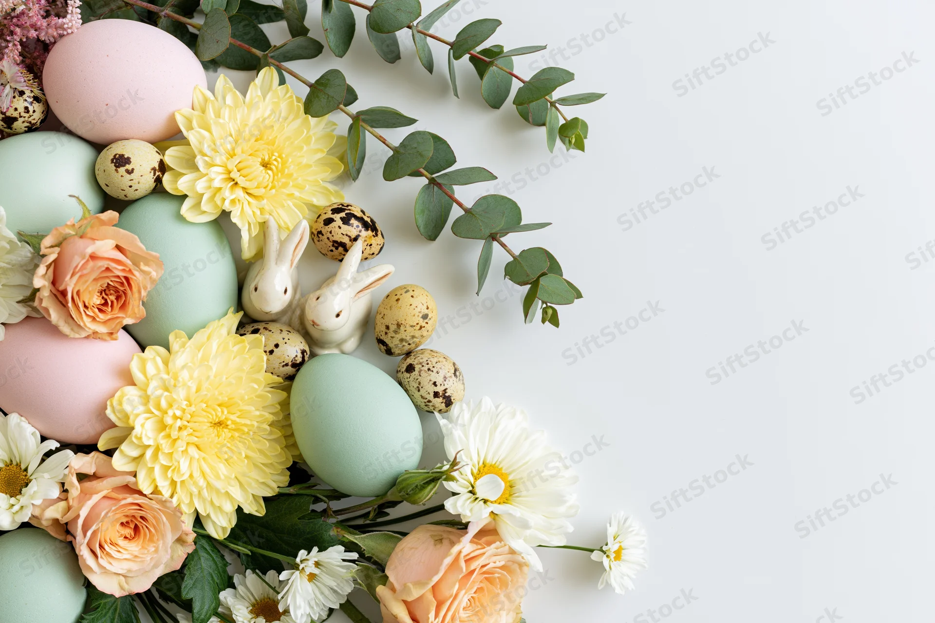 Easter flat lay arrangement with pastel eggs, flowers, and bunnies in bottom left corner on white background