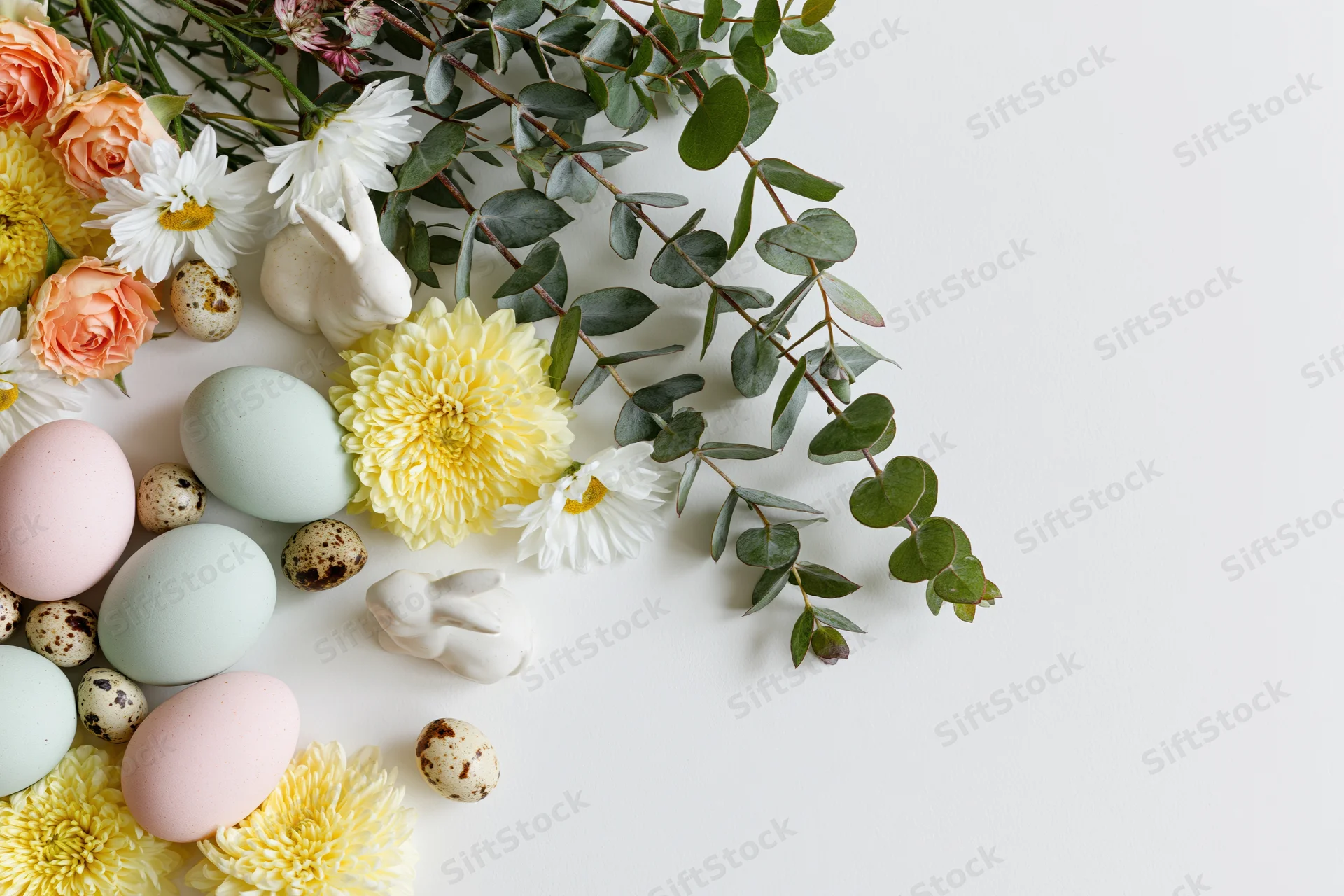 Easter flat lay arrangement with pastel eggs, flowers, and ceramic bunnies in bottom-left corner on white surface