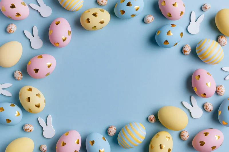 Pastel-colored easter eggs and bunny cutouts arranged in a flat lay frame on a light blue background with copyspace in the center