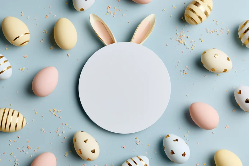 Blank white card with bunny ears surrounded by decorated easter eggs and confetti on pastel blue surface