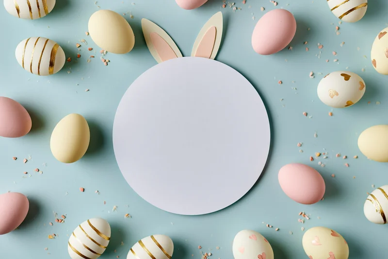 Flat lay easter scene with blank white circle card centered among decorated eggs and bunny ears on pastel blue background