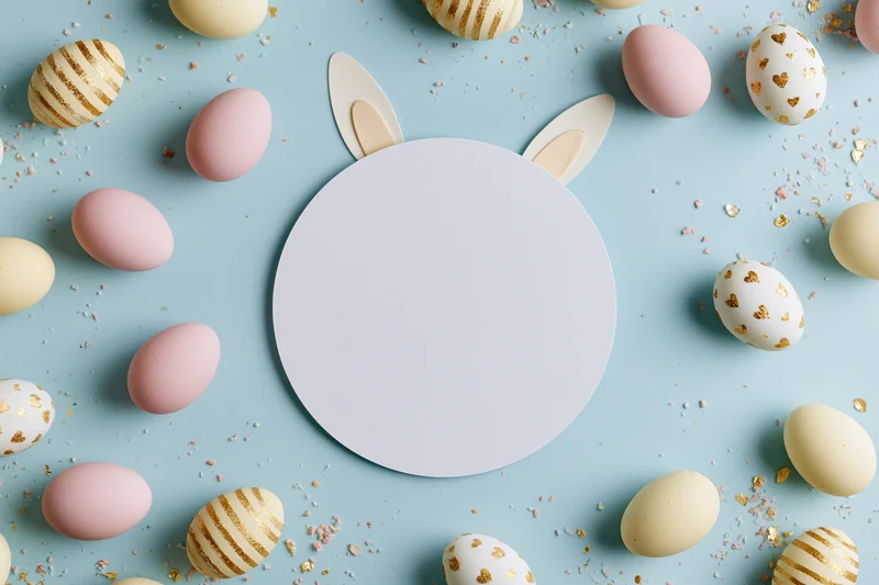 Flat lay easter scene with blank white circular card centered among decorated eggs and bunny ears on pastel blue background