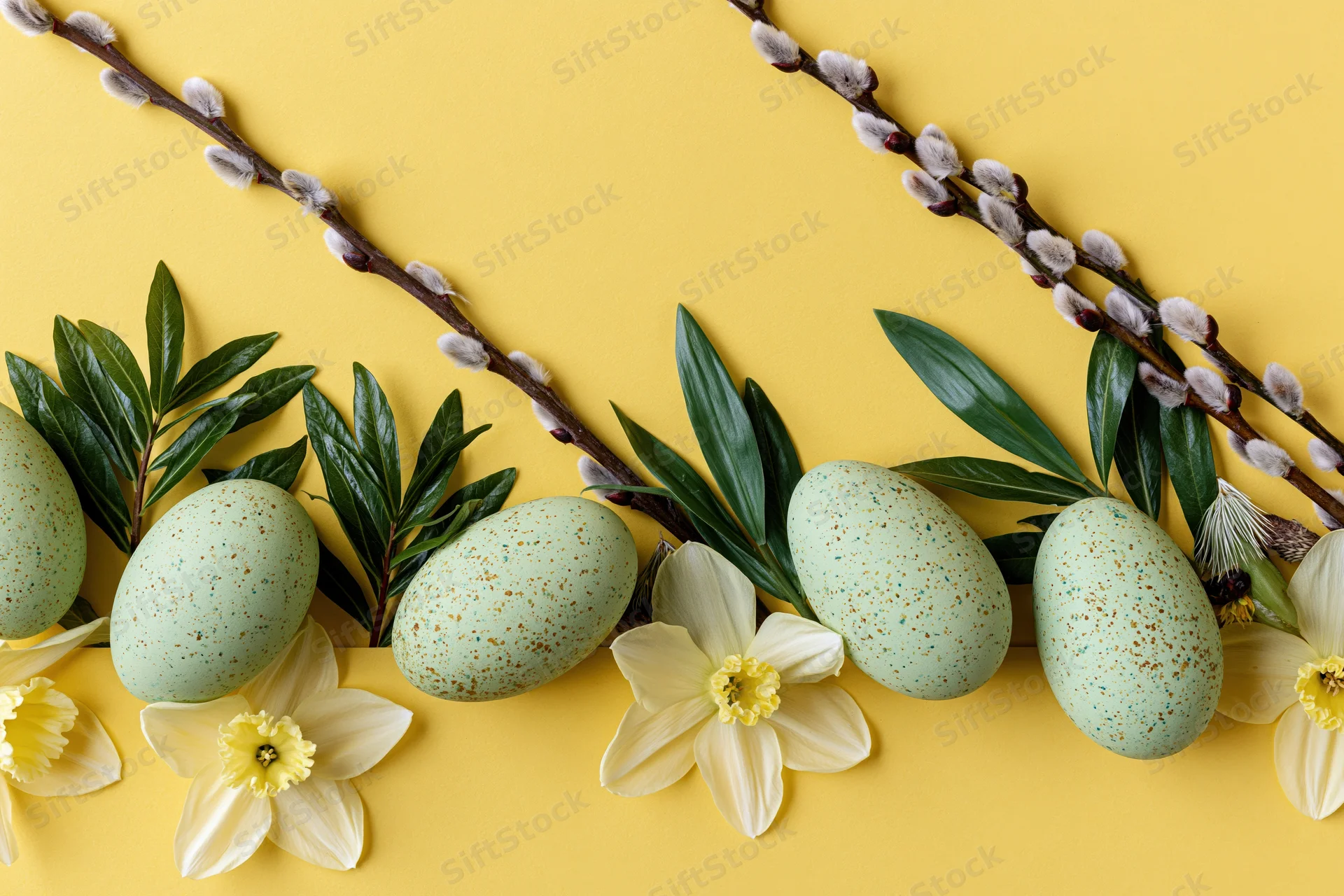 Pastel green speckled easter eggs arranged with daffodils and willow branches on yellow background