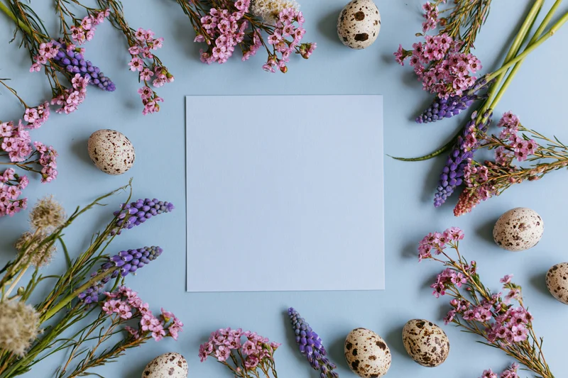 Blank pale blue square card centered on light blue surface surrounded by pink and purple flowers and speckled quail eggs in flat lay arrangement