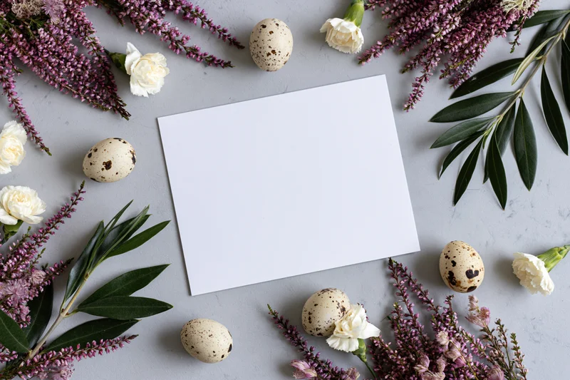 Blank white card centered on light gray surface surrounded by quail eggs, purple heather, white carnations and green leaves in flat lay arrangement