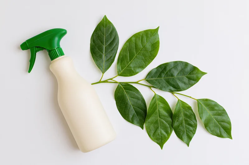 Cream-colored plastic spray bottle with green nozzle and fresh green leaves arranged in flat lay on white background