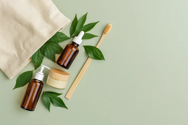 Natural skincare products and bamboo toothbrush arranged with green leaves on a flat surface beside a canvas tote bag
