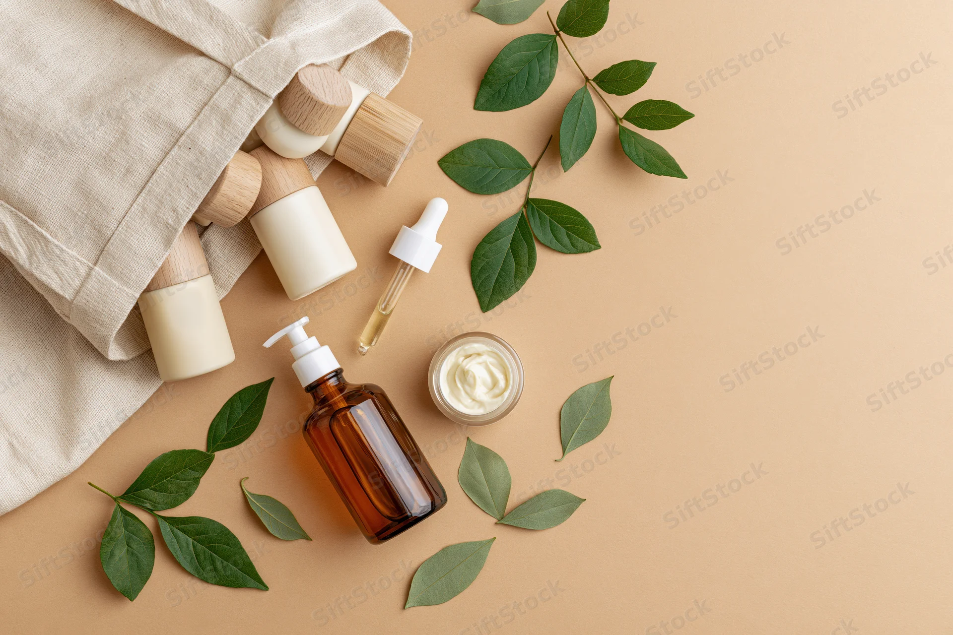 Cream canvas tote bag with wooden-capped skincare bottles and green leaves arranged in flat lay on beige surface