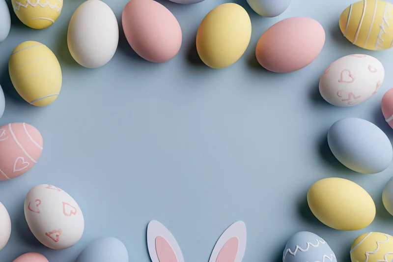 Decorated easter eggs arranged in a frame on a pastel blue surface with bunny ears and copyspace in center