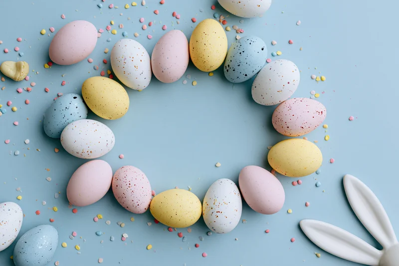 Decorated easter eggs arranged in a circular frame on a pastel blue background with confetti and bunny ears