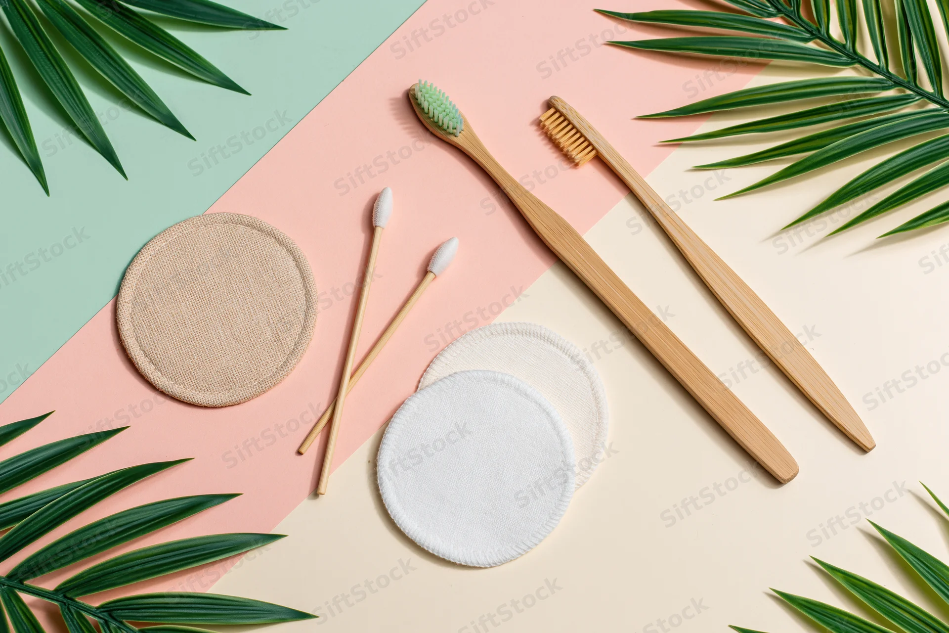 Bamboo toothbrushes and reusable cotton pads arranged in flat lay with palm leaves on pastel background